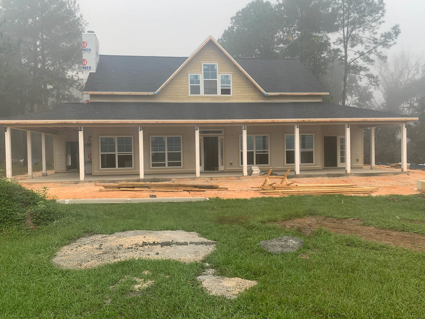 Wood-framed house under construction with exposed roof, surrounded by trees and patches of grass, dirt, and scattered stones