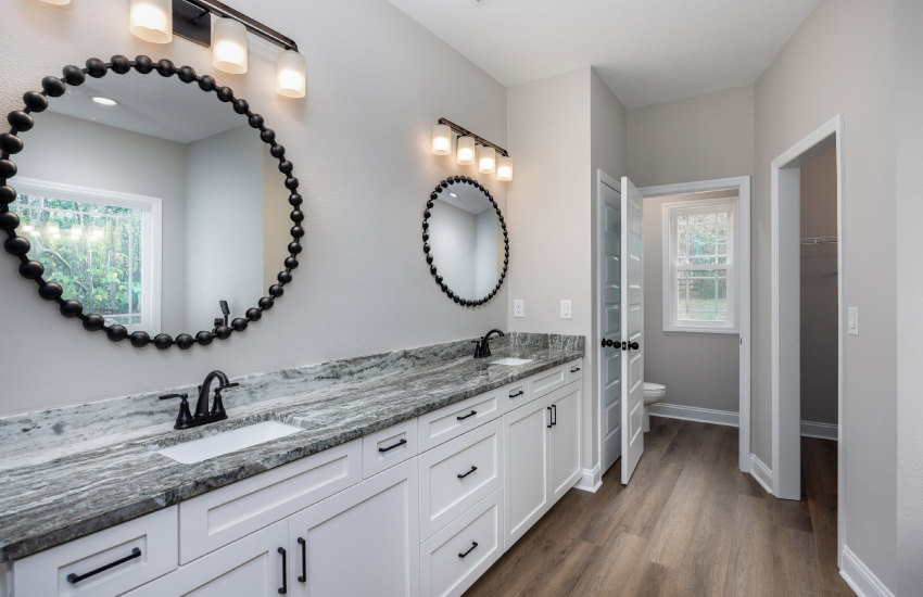 White bathroom cabinets with marble countertops, rectangular mirror above sink, row of wall-mounted lights, black faucet, window with black bead border, tile flooring.