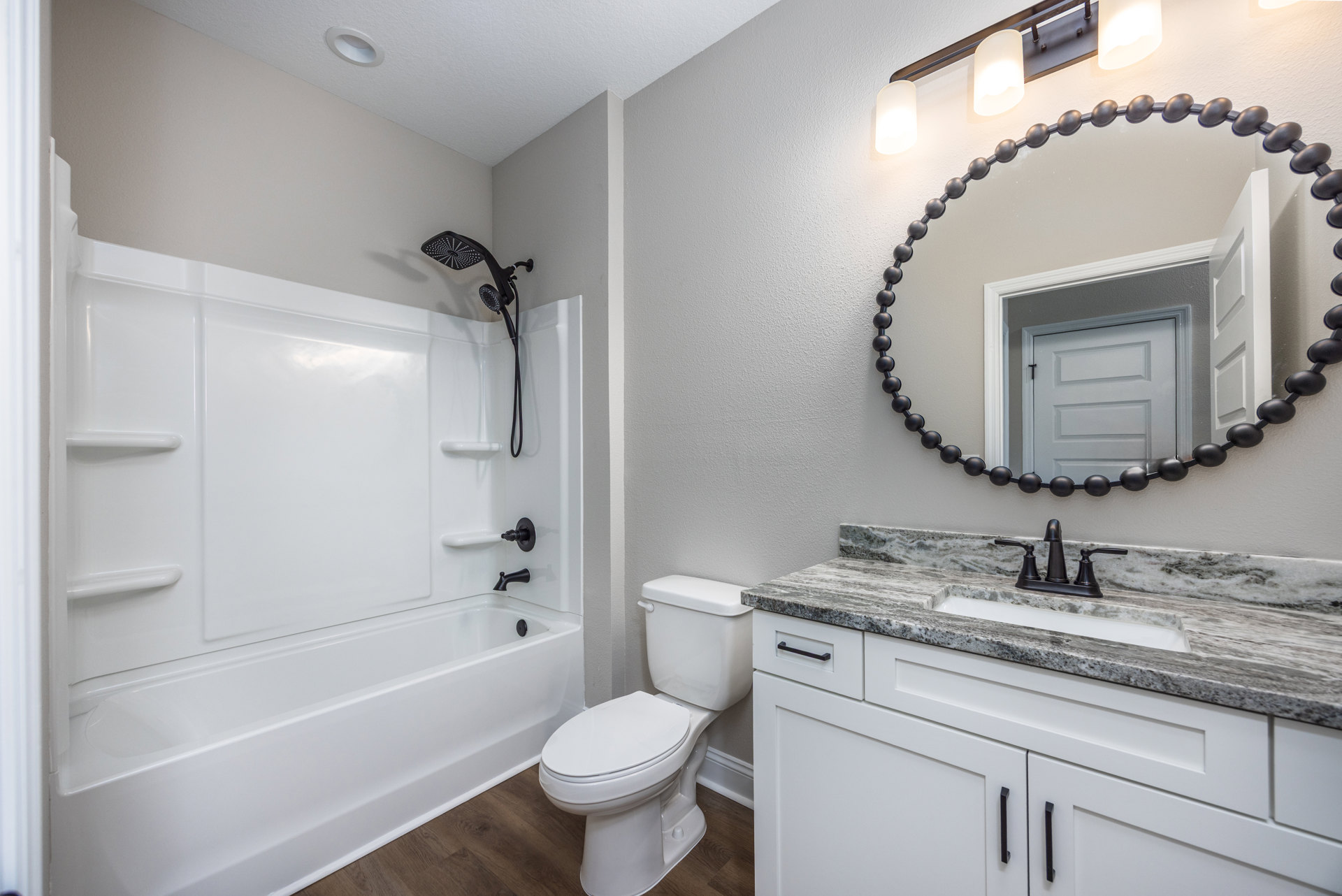 Bathroom with white toilet, freestanding bathtub, wall-mounted shower head with hose, large mirror above vanity, white door featuring black round knobs, light tile flooring and