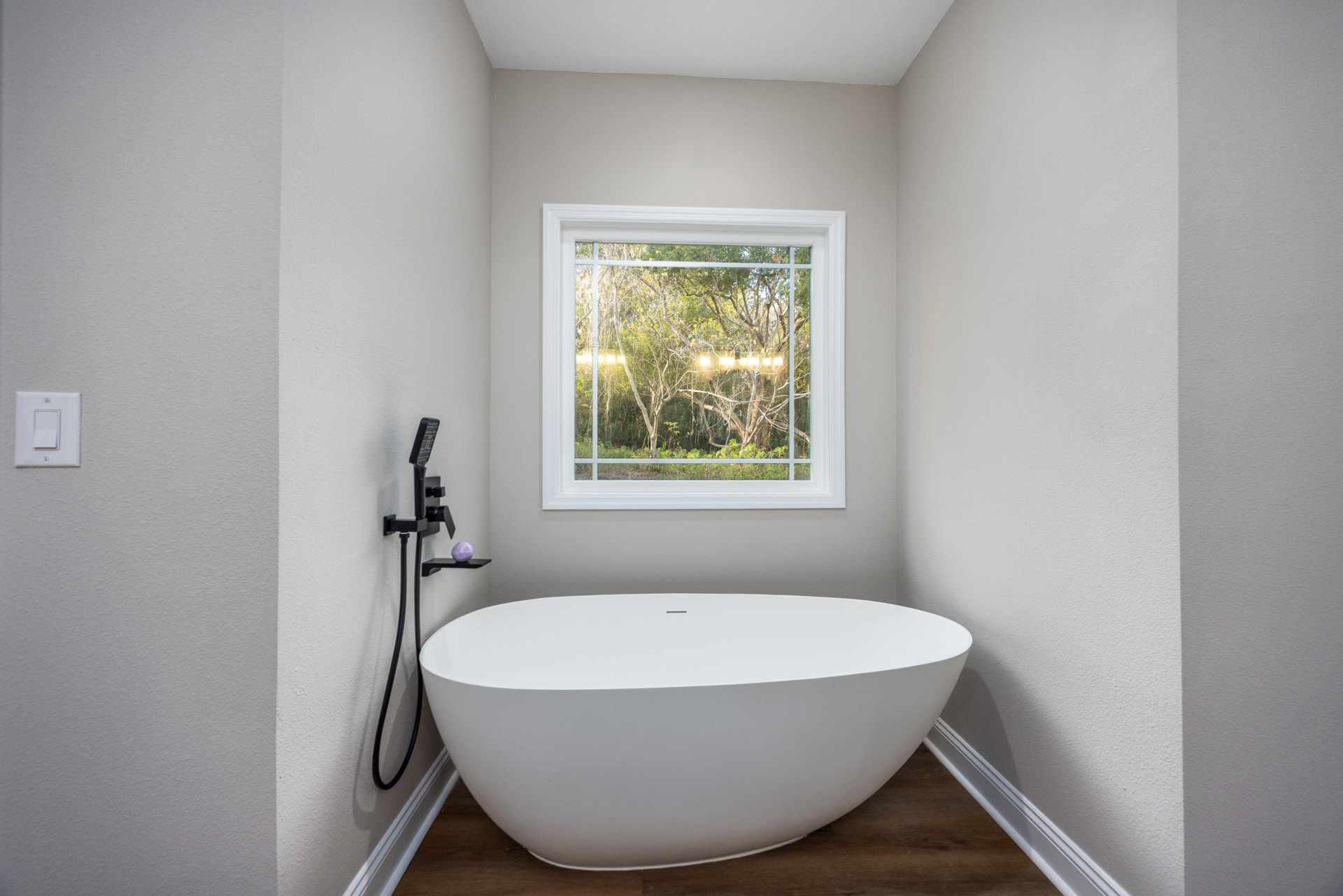 White oval bathtub beside a black shelf and white sink, large window with trees outside, light switch on tiled wall