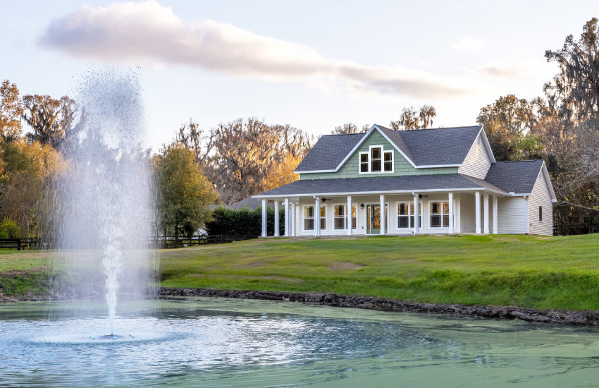 Two-story house with green roof, large windows, and covered porch, landscaped front yard featuring stone water fountain surrounded by grass, trees, and flowering plants under