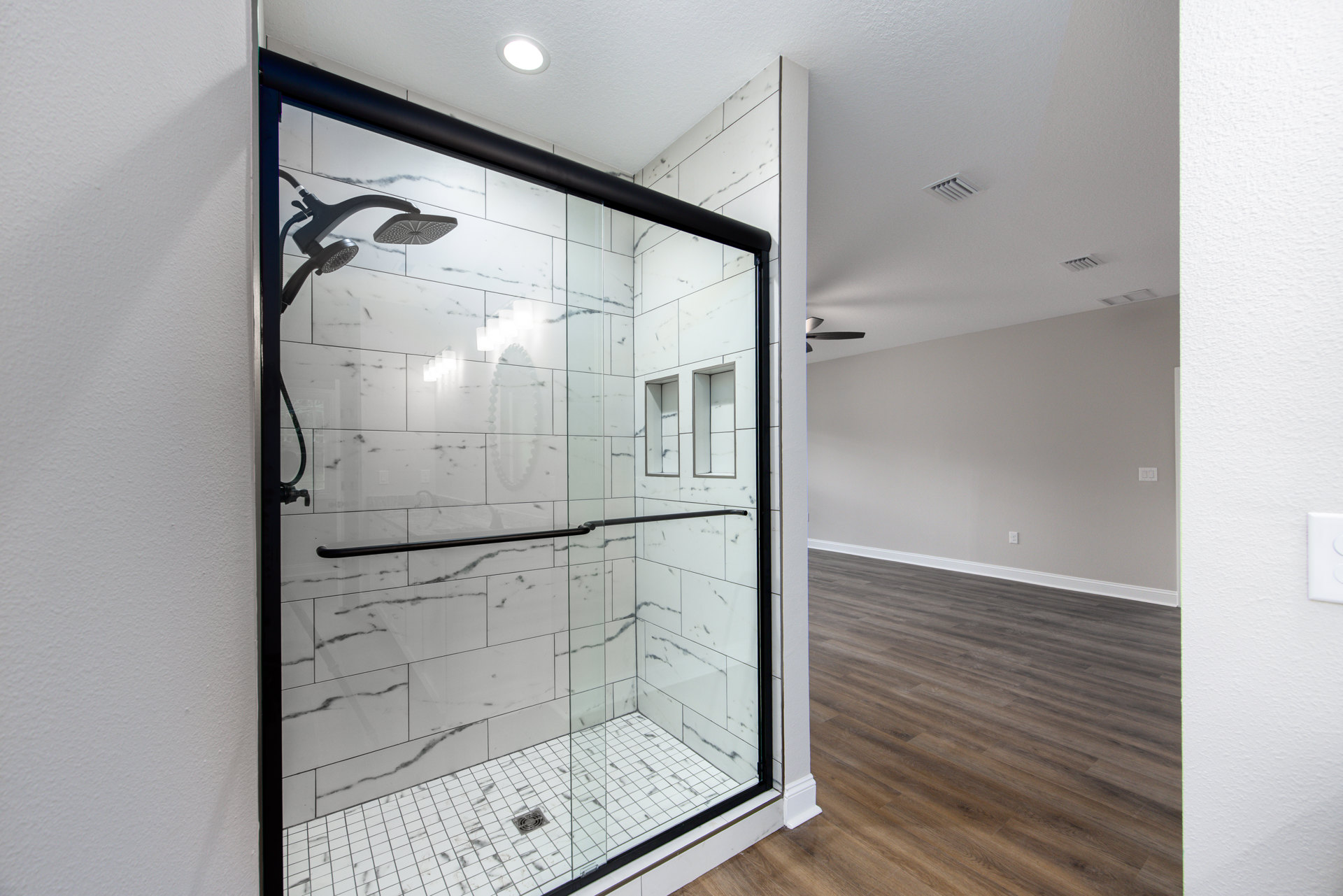 Glass shower enclosure with black hardware, white tile walls, ceiling light, and floor drain
