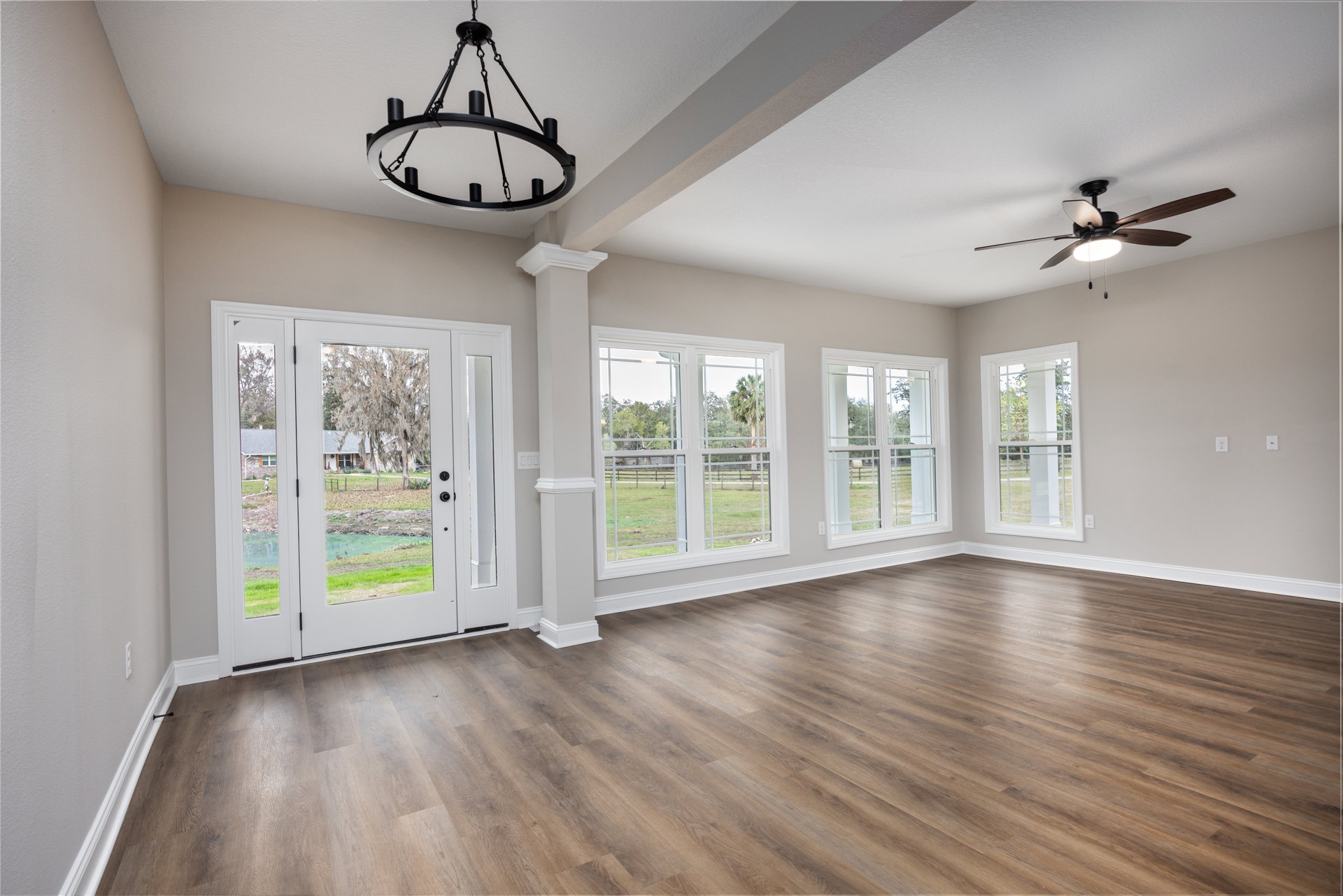 Hardwood floor room with white walls, black candle-style chandelier, ceiling fan with light, large window framed in white overlooking fenced grassy yard