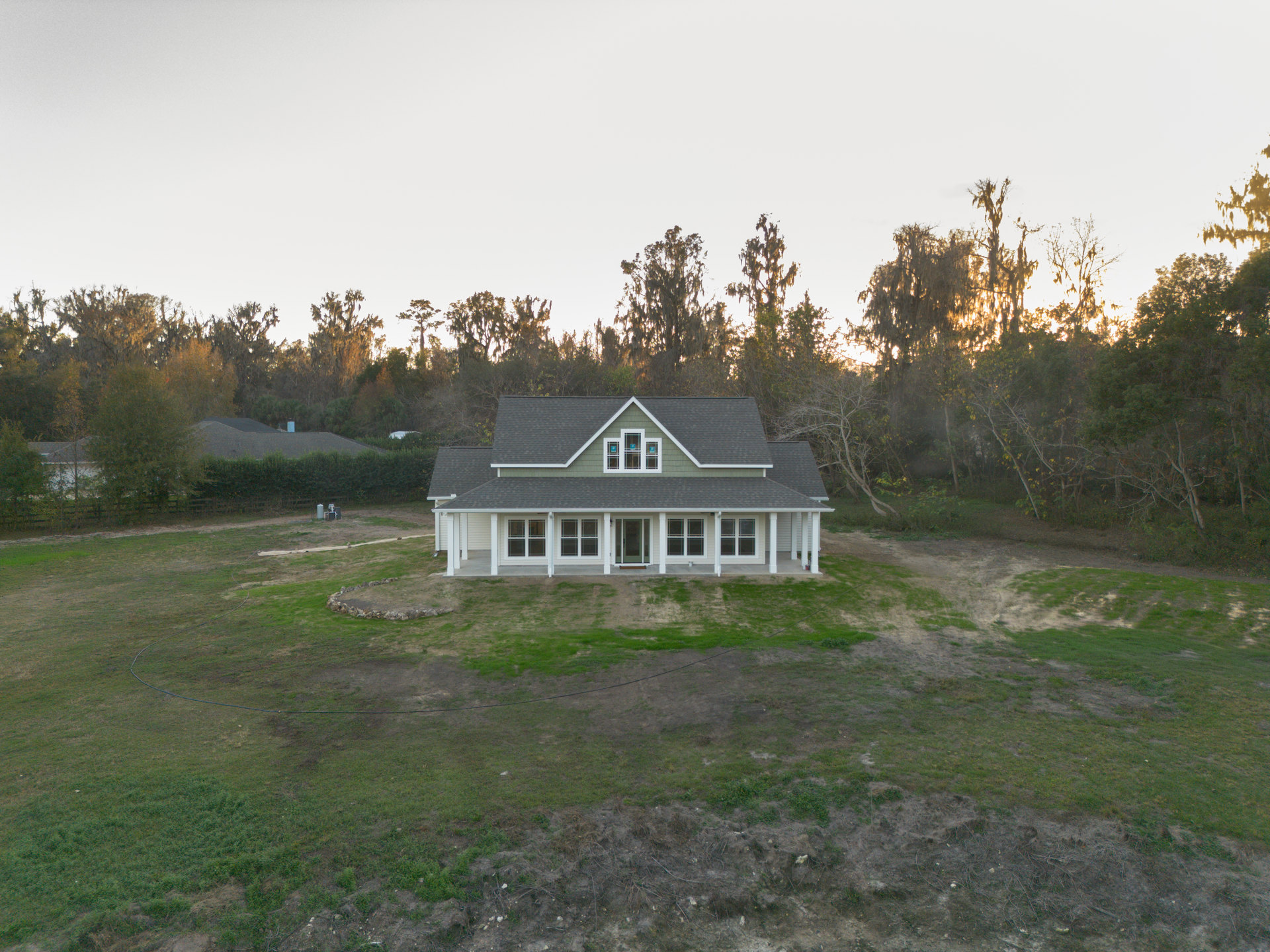 Two-story farmhouse with green roof, white siding, expansive lawn, mature trees, and large windows under clear blue sky