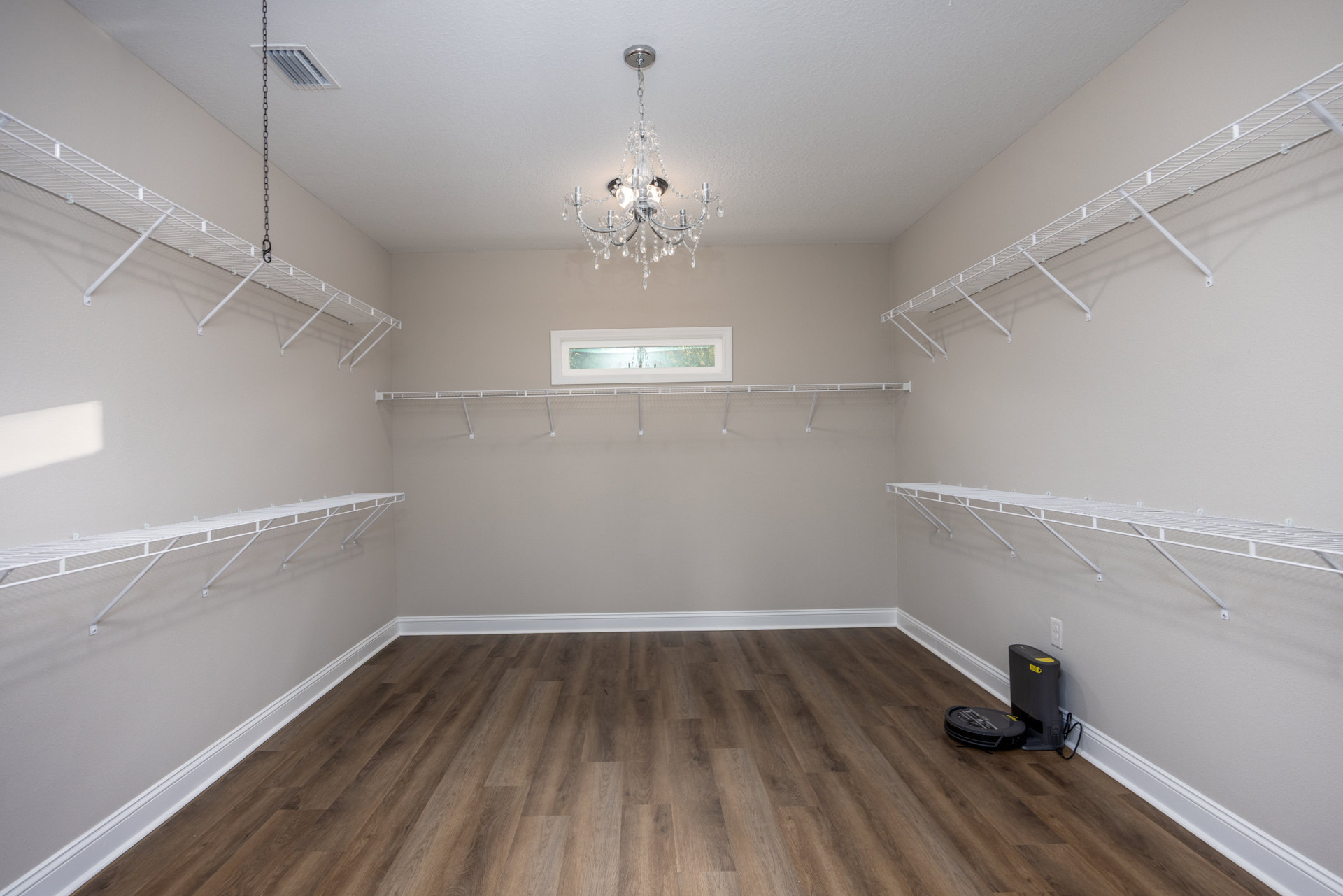 White walls with built-in shelves, wood flooring with white trim, chandelier hanging from ceiling, window close-up, vacuum and black box with yellow label placed near shelving.