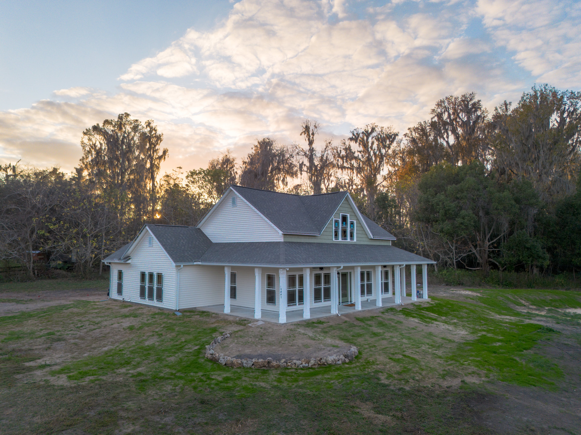 Wide front porch with white columns overlooking expansive green lawn, stone circle feature near entry, rectangular windows, gabled roof, mature trees and partly cloudy sky in