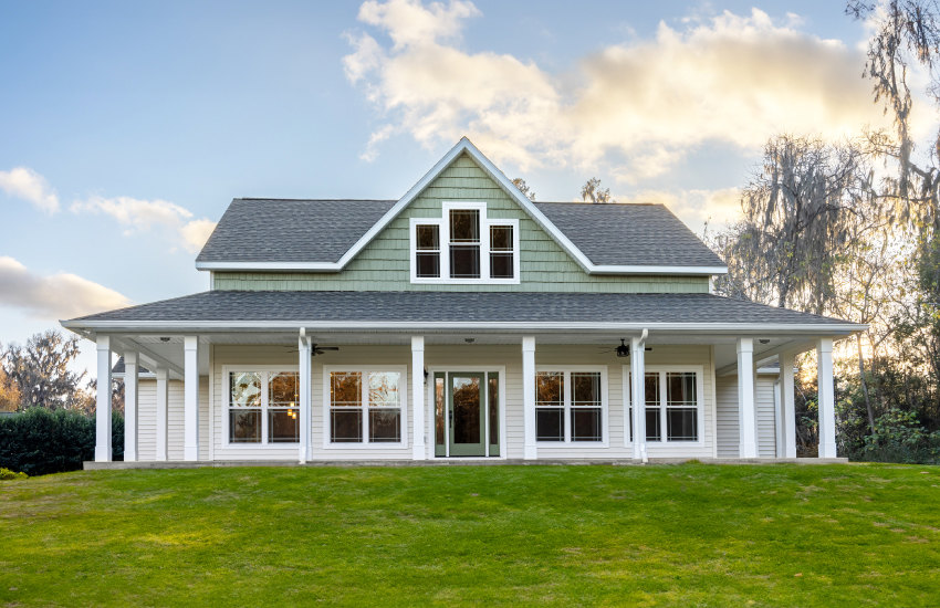 White house with triangular green roof, large front window, and wooden front door, surrounded by a manicured green lawn under a partly cloudy sky