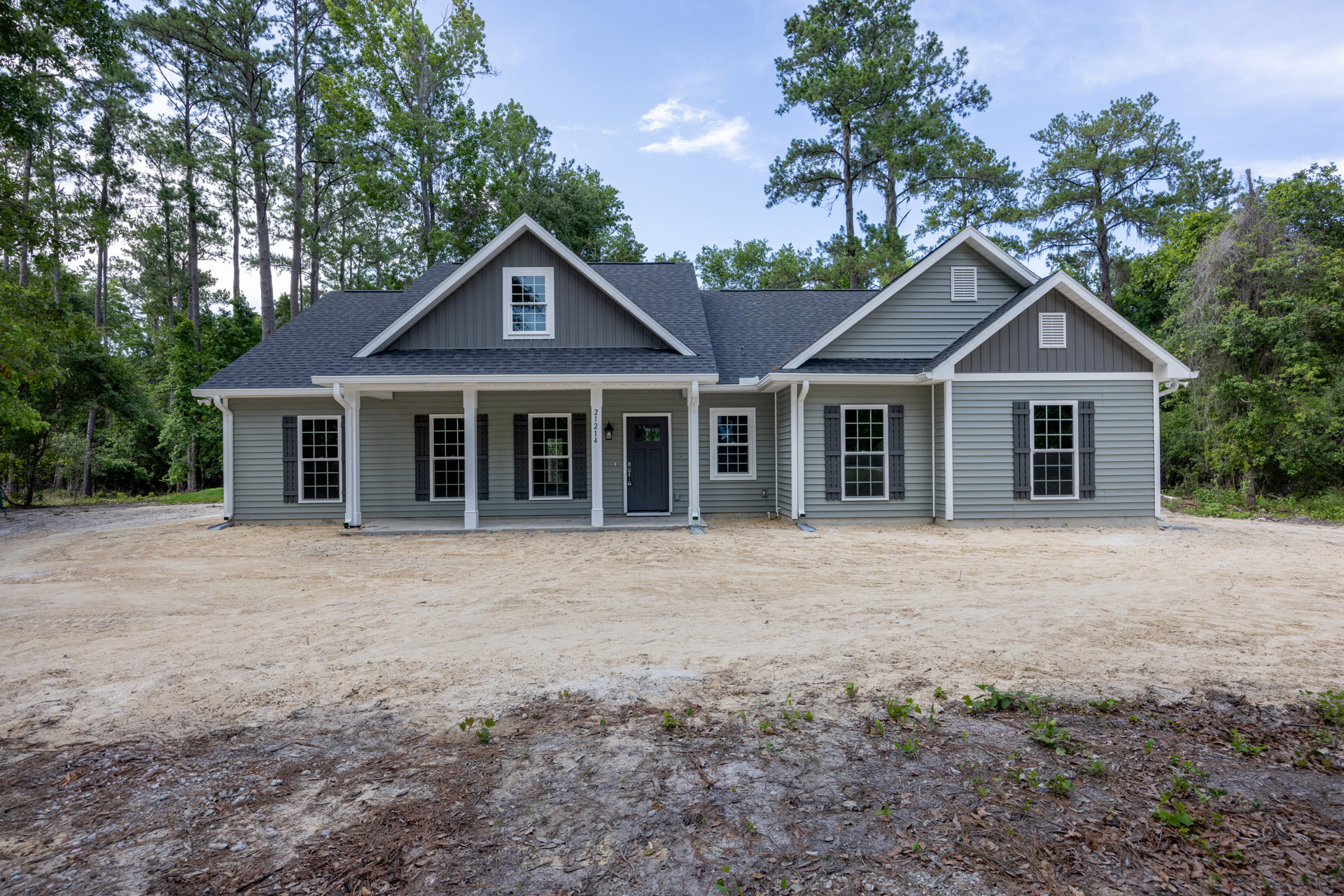 White house with grey roof, black front door with lock, white-trimmed windows, dirt road and patch in front, trees and cloudy sky in background