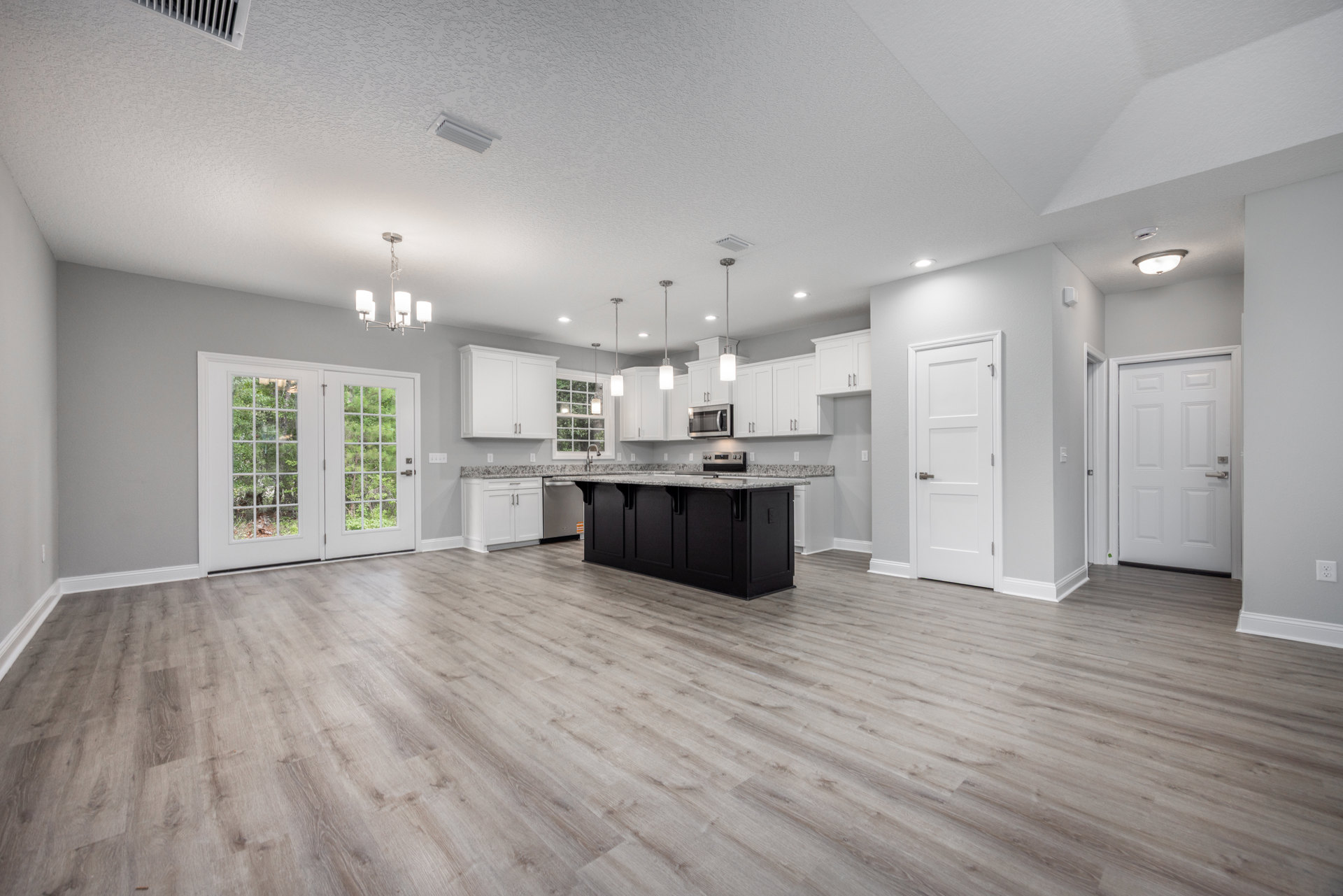 Spacious kitchen with white cabinets, grey accents, black island topped with marble, wood flooring, double doors opening to tree views, and silver-handled white door.