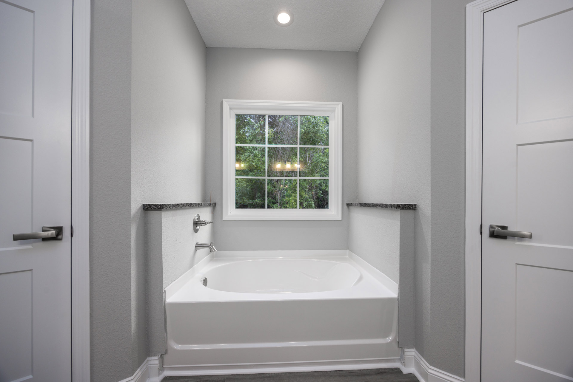 Freestanding white bathtub beneath large window with tree views, white tile walls, ceiling light fixture, silver-handled door, modern sink and tap