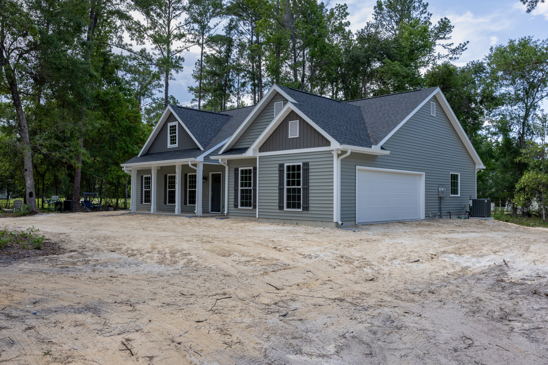 Partially built house with white-framed windows, white garage door, gray roof, dirt yard, and construction equipment, surrounded by trees in the background