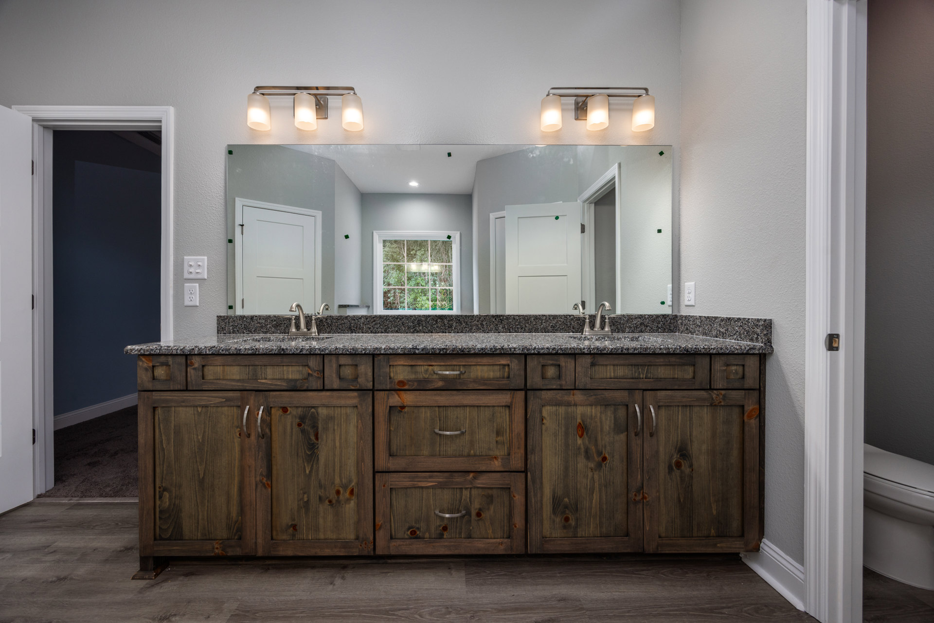 Bathroom with expansive mirror above double sinks, quartz countertop, white cabinetry, modern light fixture, window revealing leafy trees, and white paneled door with silver
