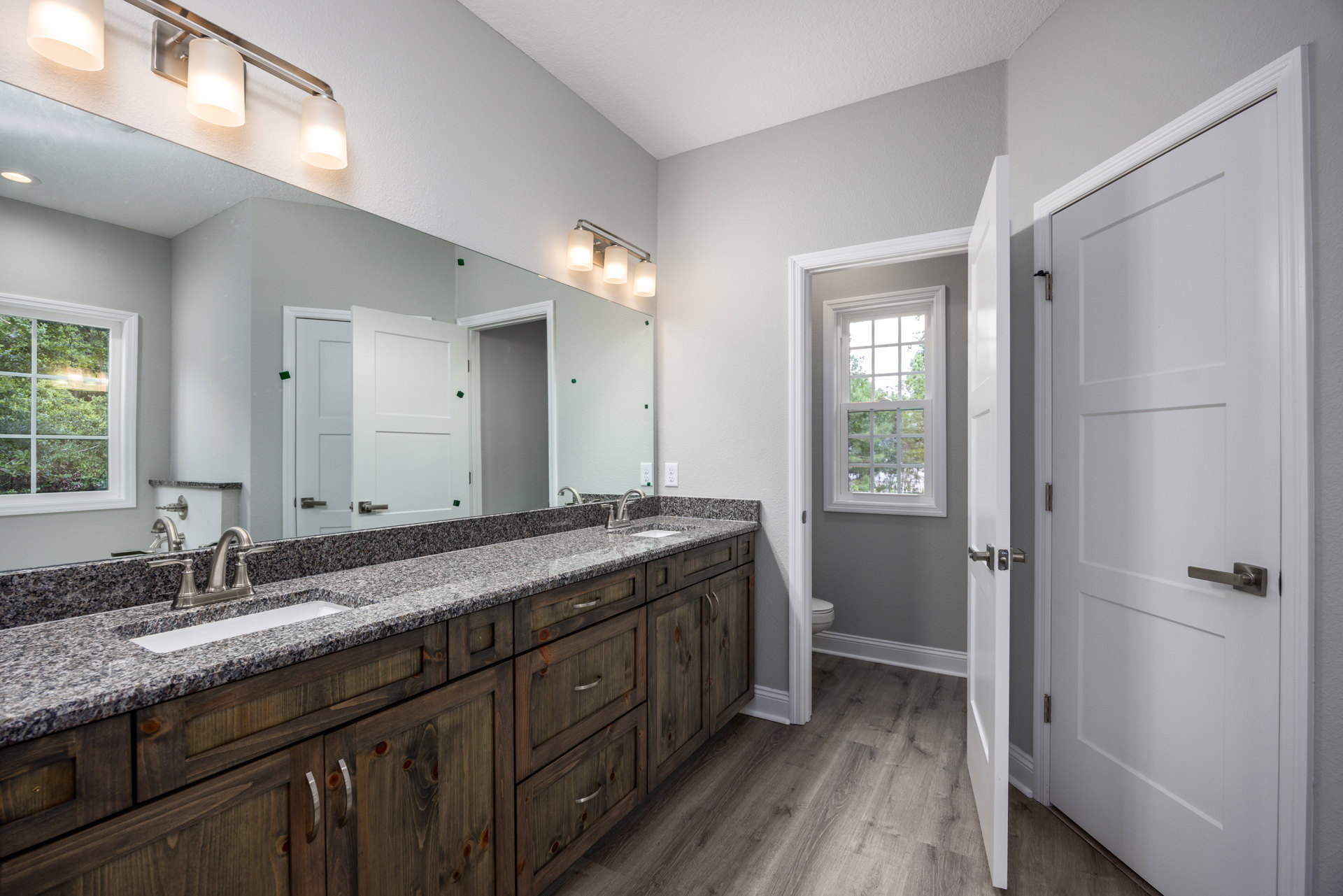 Bathroom with double white sinks, wide mirror above, white-framed window showing trees outside, light gray tile backsplash, modern chrome faucets, and contemporary light fixtures.