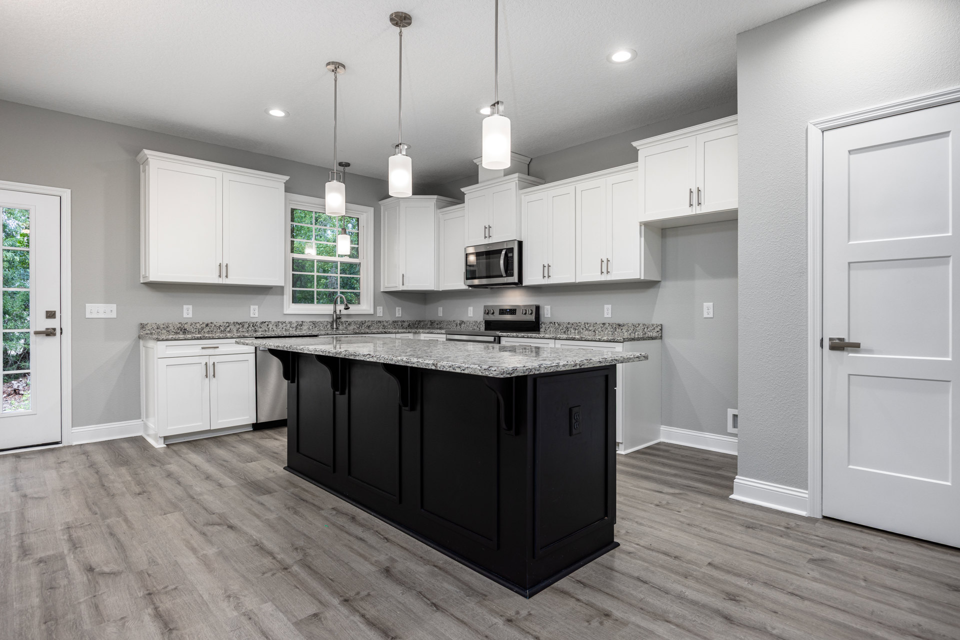 Marble kitchen island with black base, white cabinets, tile backsplash, stainless steel microwave, white doors with silver handles, light fixture above island, wood flooring