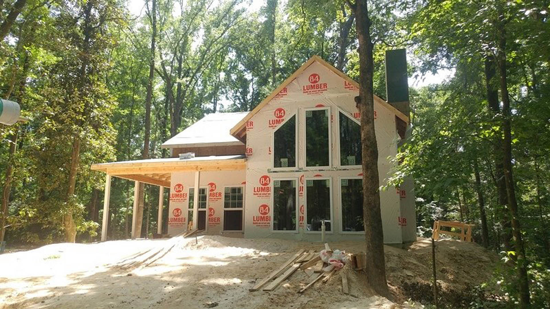 Wood-framed house under construction surrounded by tall trees, unfinished porch, exposed windows with multiple panes, pile of lumber on dirt ground