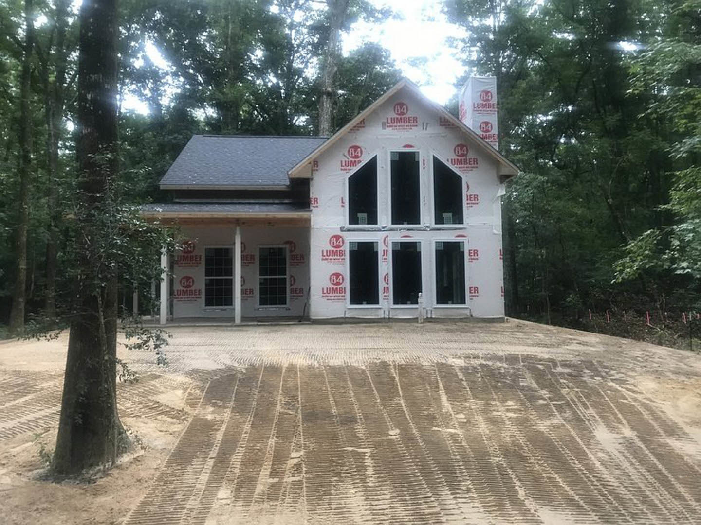 Wood-framed house under construction with installed windows, surrounded by tall trees and dirt ground with tire tracks, partially visible porch and roof, black rectangular