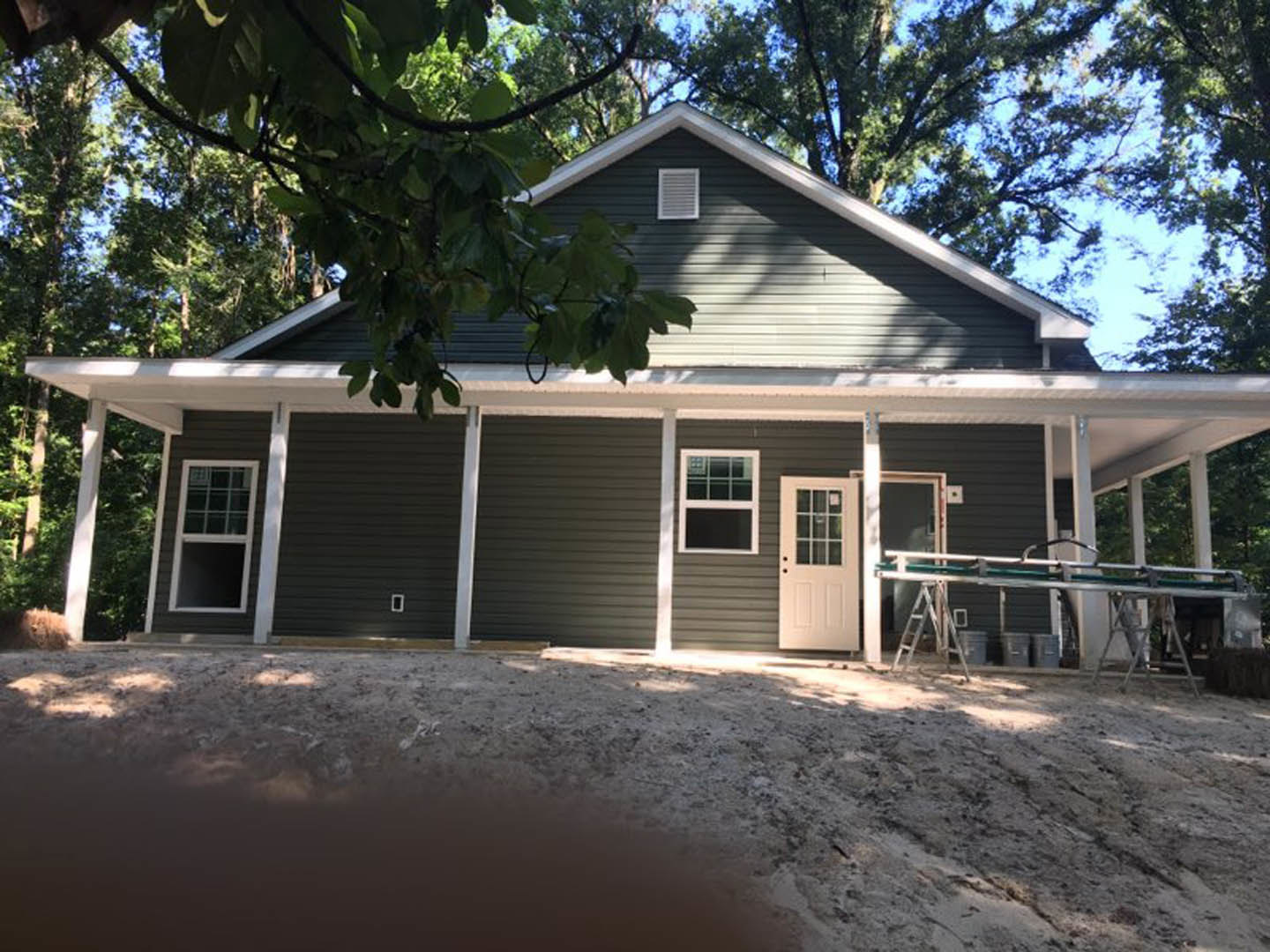 Framed house under construction with exposed plywood siding, white door with glass panes, square window, wall vent, metal structure with green mat, dirt ground, and trees in the