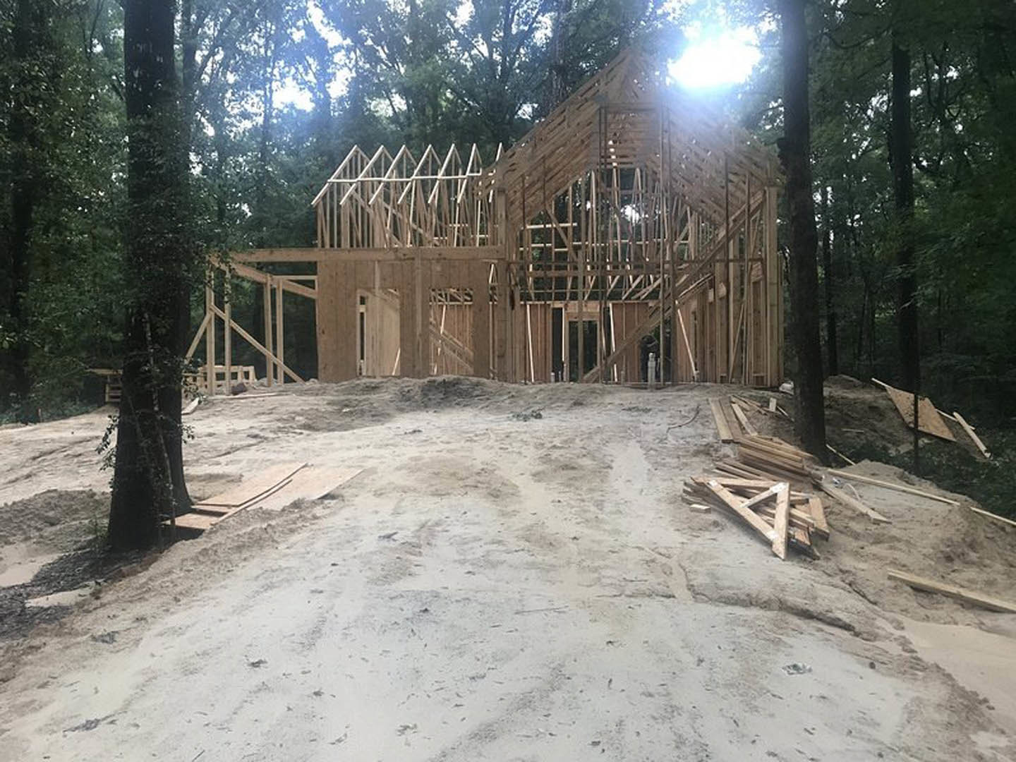 Partially built wooden house framed with exposed lumber, surrounded by tall green trees, dirt piles, and stacks of wood planks on the construction site