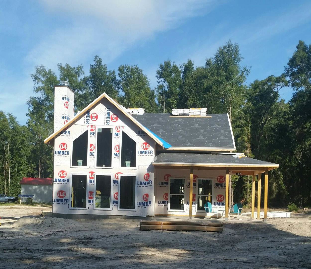 Wood-framed house under construction with exposed beams, unfinished roof, dirt ground scattered with lumber, trees and cloudy sky in background