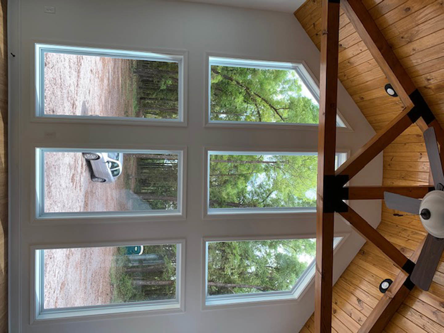 Vaulted ceiling with exposed wood beams, ceiling fan, large windows revealing parked car and trees outside, white walls, natural daylight illuminating interior.