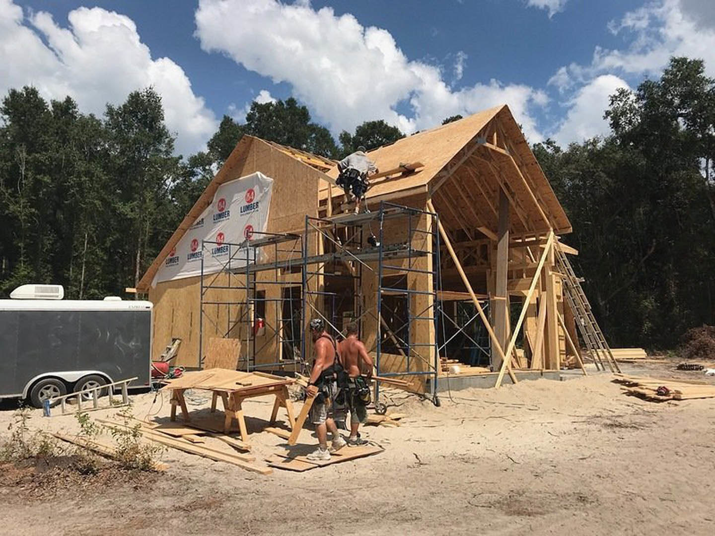 Construction workers in safety harnesses assembling wooden framing and roof structure, white tarp draped over roof, trailer parked nearby, scattered lumber and tools on ground