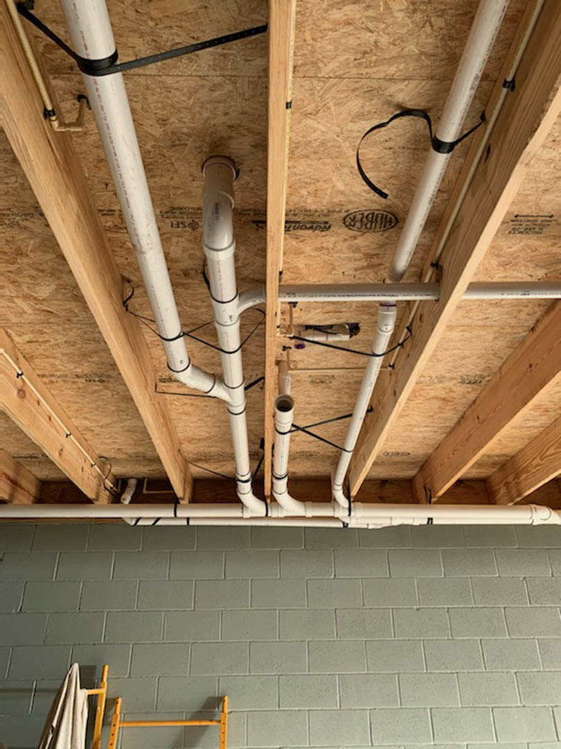 Exposed metal pipes running along a wood beam ceiling, brick wall with a yellow ladder leaning against it, towel hanging on a rack, white wall with scattered black spots, white