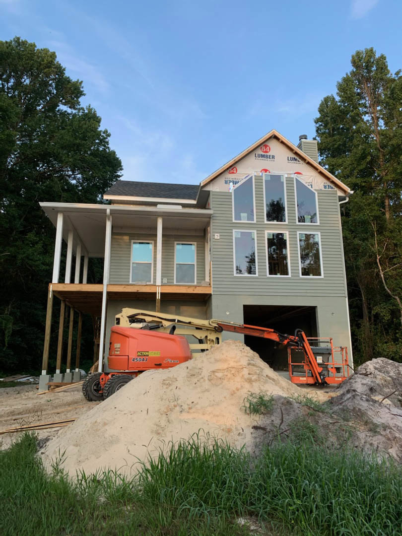 Two-story house under construction with exposed framing, pile of dirt in front yard, crane positioned nearby, large windows, trees in background, cloudy sky overhead