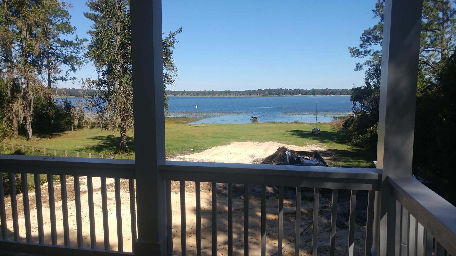 Wooden deck with metal railing overlooking a sandy beach and calm lake, trees lining the far shore under a clear blue sky, grassy area in foreground, trash can near the fence