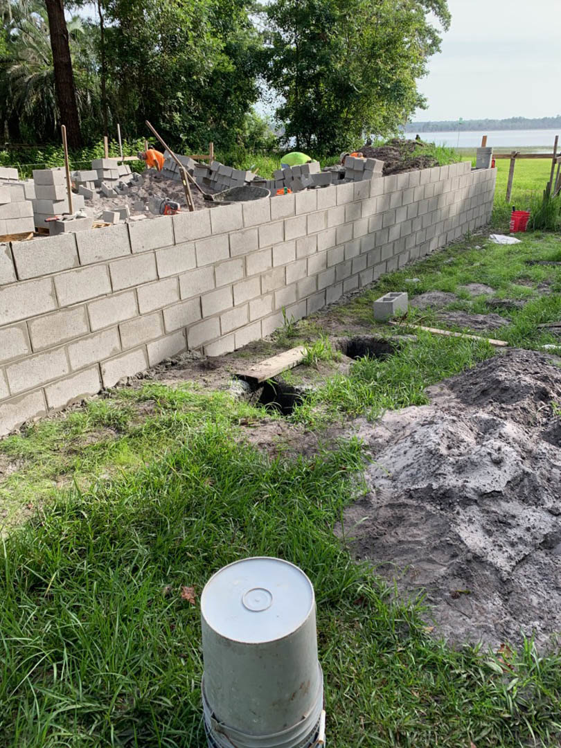 Bricks stacked beside a dirt mound and grey bucket on grassy construction site, unfinished wall with hole in ground, trees in background