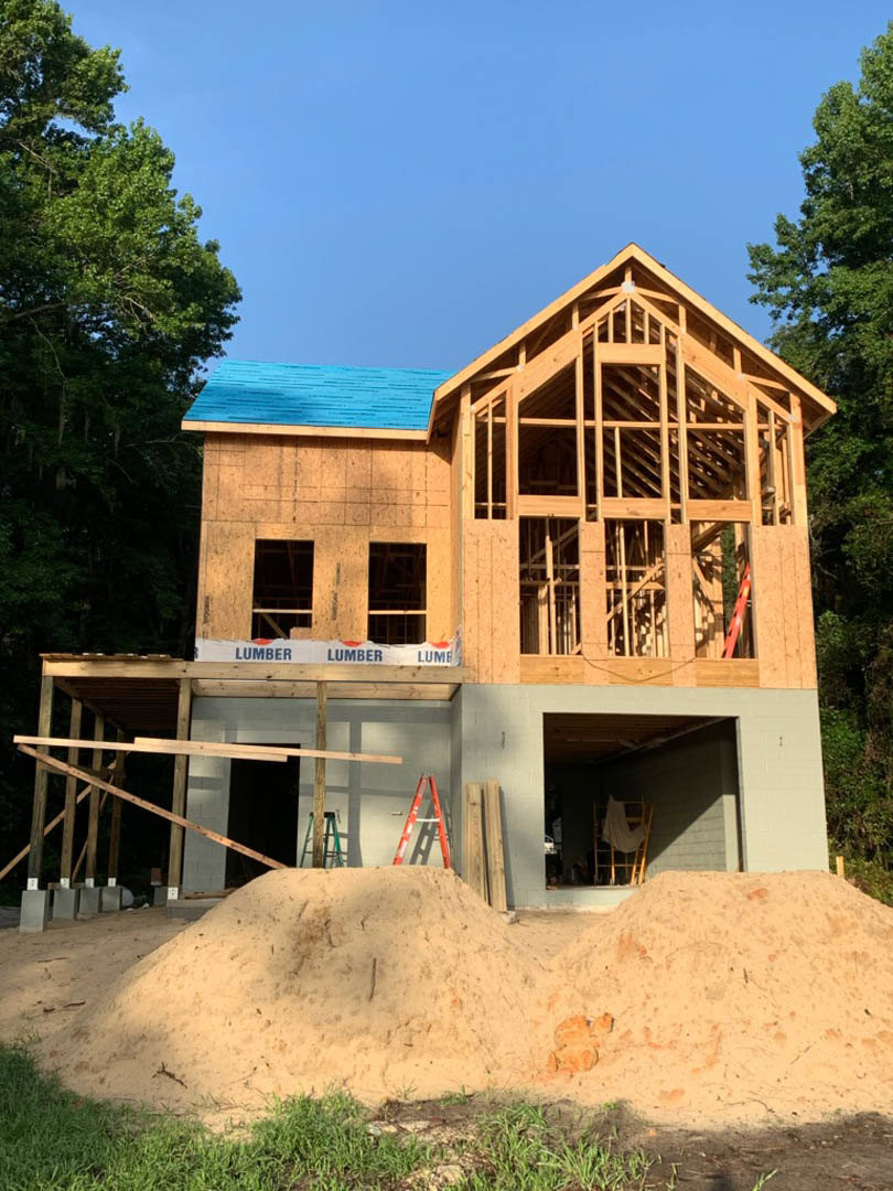 Partially built house with blue roof, exposed lumber framing, sand pile in foreground, surrounding trees, and construction materials scattered outside
