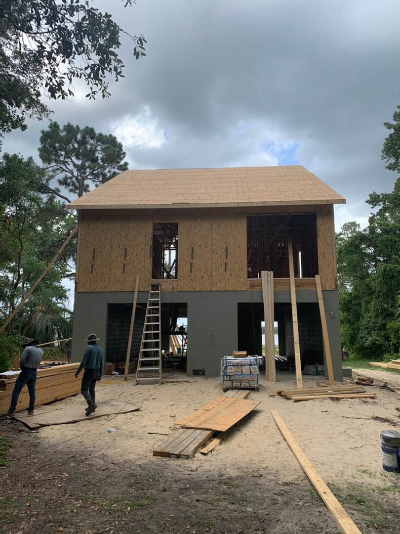 Framed house under construction with exposed lumber, ladder leaning against exterior, people walking on dirt ground, cloudy sky overhead, trees in background