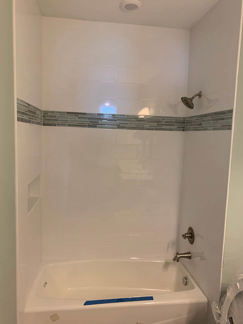 White-tiled shower with chrome shower head and faucet, partial view of bathtub, plastic bag resting on toilet, wall-mounted mirror above sink