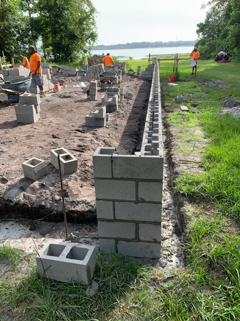 Group of people standing beside a stacked concrete block wall outdoors, with grass, soil, and trees visible in the background