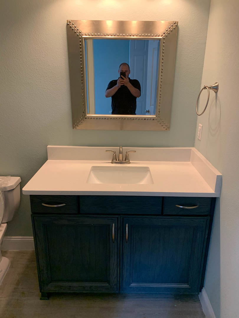 White bathroom with tile walls, chrome towel holder, white countertop, black soap dispenser, large mirror reflecting man taking a selfie