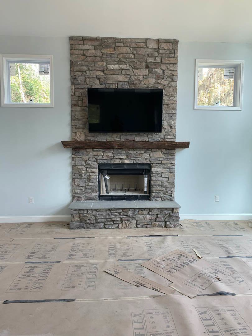 Stone fireplace with mounted flat-screen TV above, white-framed window to the side, papers scattered on hardwood floor, neutral walls and hearth in modern living room.