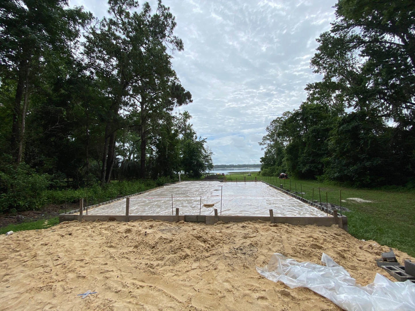 Partially constructed backyard pool surrounded by sand, mature trees, and cloudy sky, with construction materials visible near the water’s edge