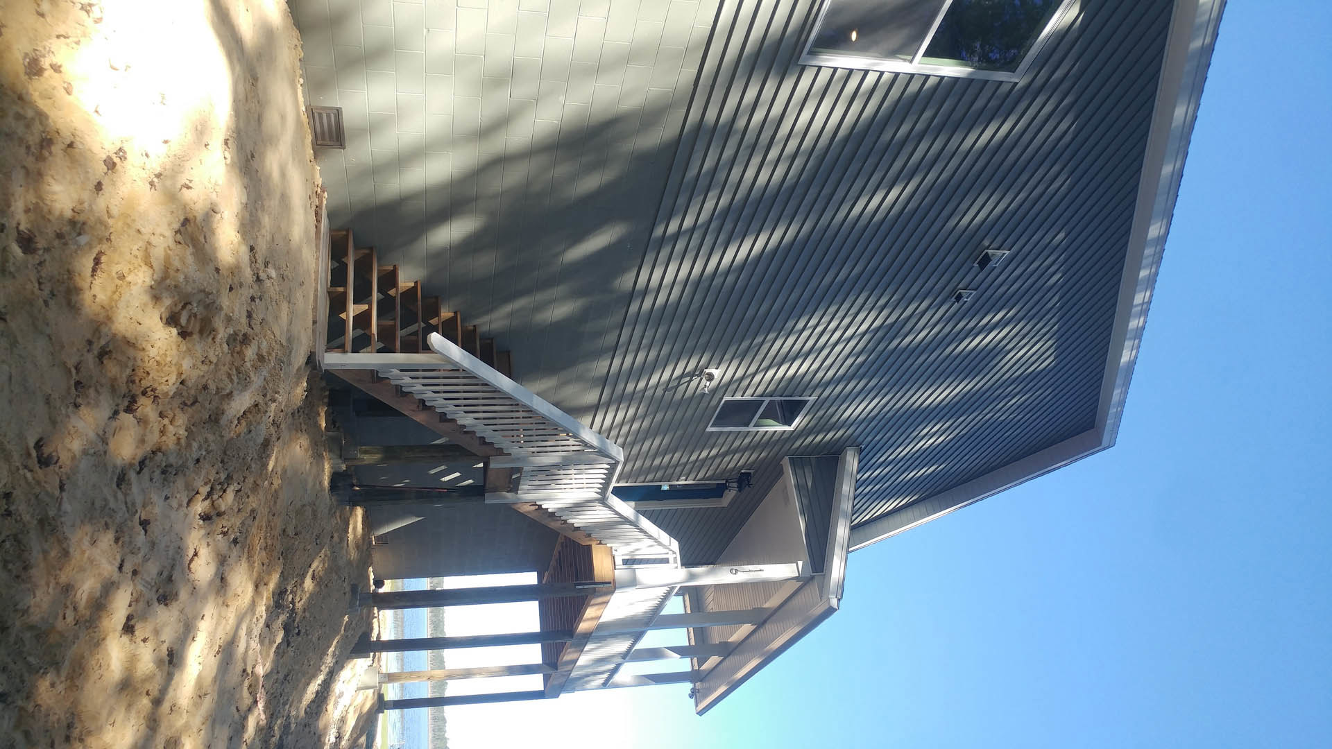 White composite front porch with exterior staircase, wooden door, and dirt road bordered by a white fence under a clear sky