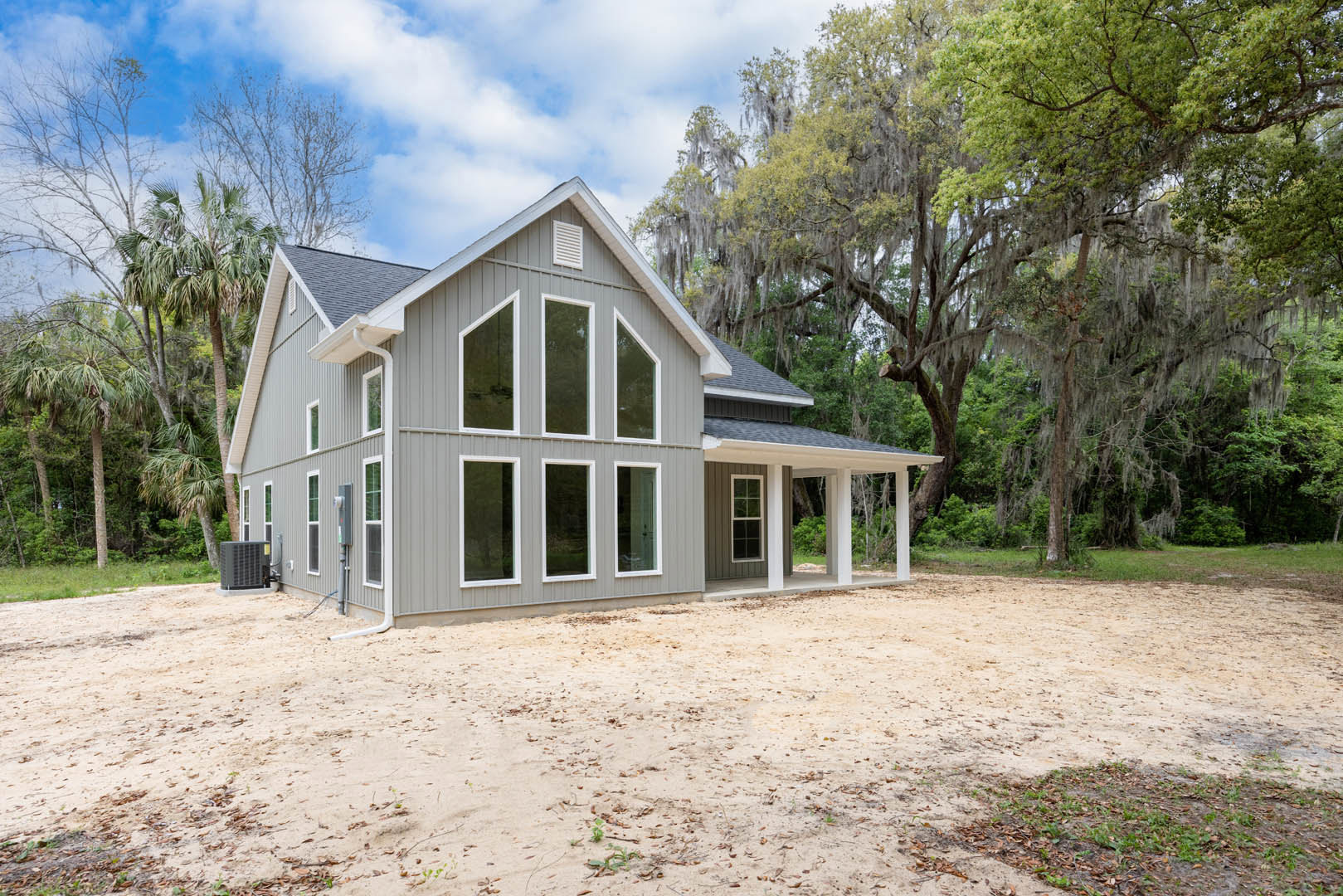 Grey siding and white trim exterior with large windows, white-framed door, covered porch, dirt ground scattered with leaves, mature trees in the background, cloudy sky overhead