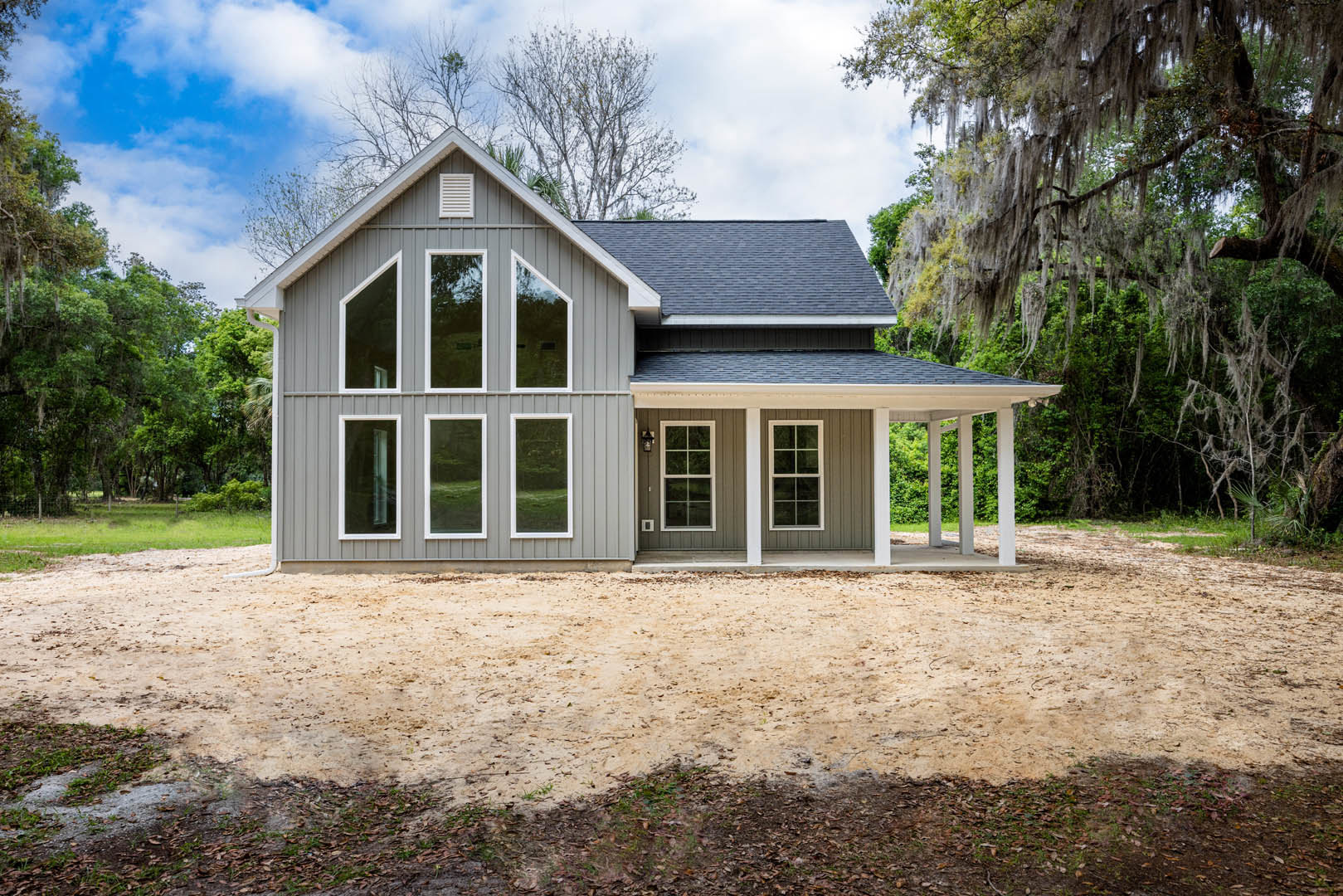 Spacious front porch with white columns, multiple large windows with white frames, gray shingle roof, white wall vent, dirt and leaf-covered ground, surrounded by trees under a