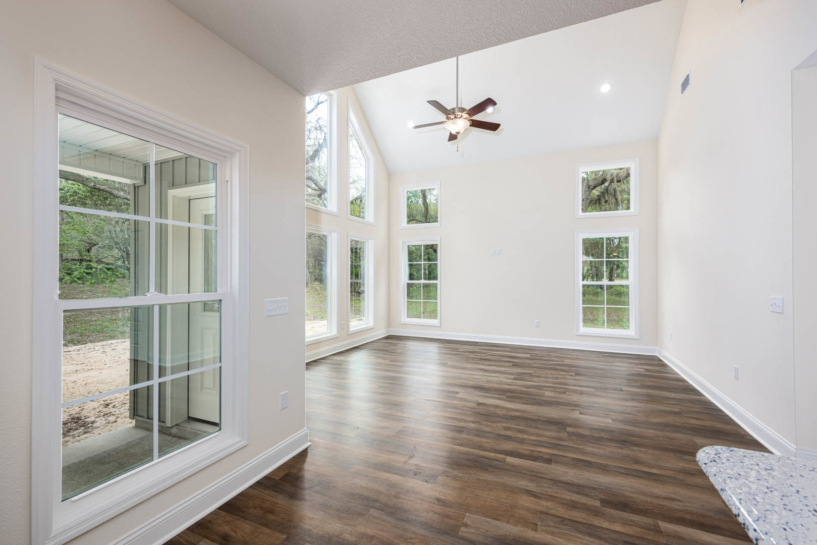 Wood flooring in a bright room with large windows overlooking trees and a dirt path, ceiling fan with light fixture, marble table in foreground