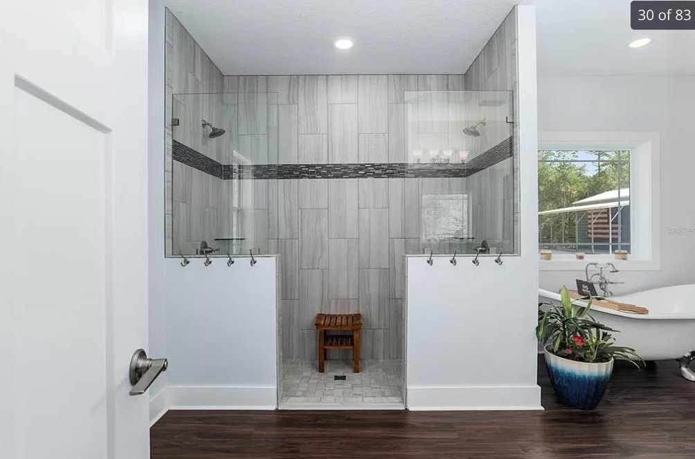 Bathroom with wood flooring, glass-enclosed shower featuring a built-in bench, potted plant beside a wooden table with lower shelf, white wall with hooks, and close-up of a