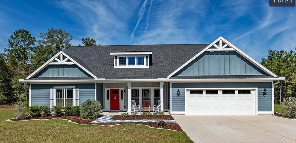 Two-story house with white siding, red front door, white trim, attached garage with windowed door, concrete driveway, row of green bushes along the front, cloudy sky overhead.