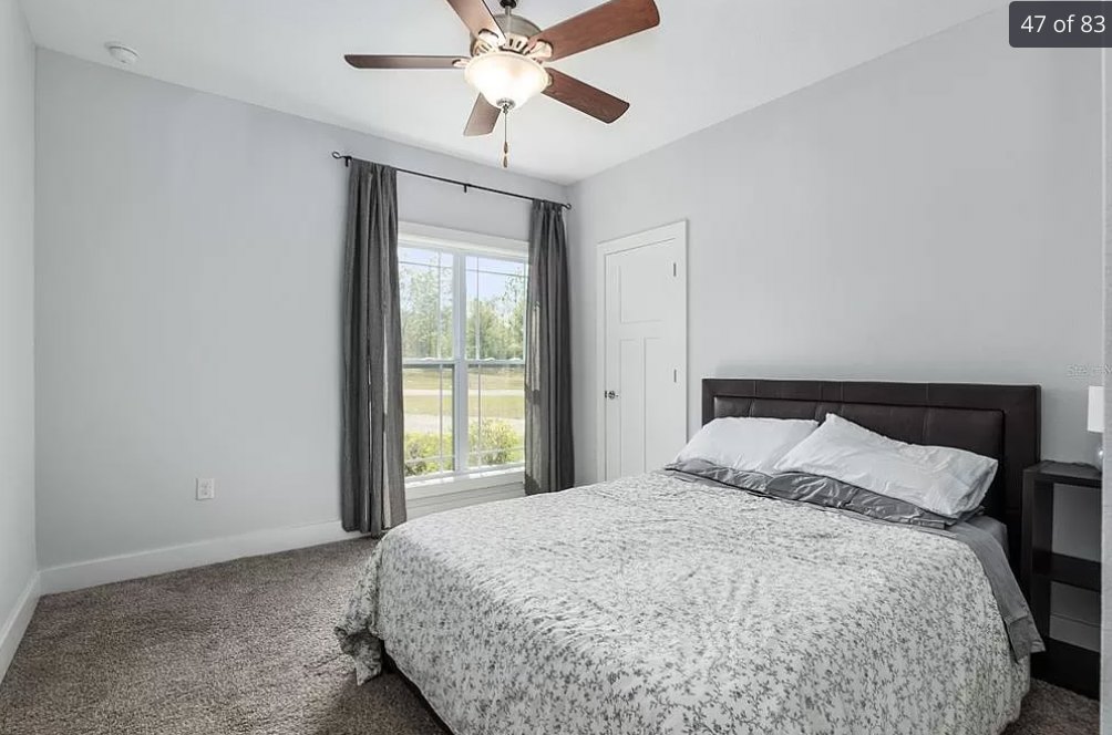 Bedroom with white bedding and pillows, ceiling fan with light fixture above, window with curtains showing outdoor view, white door with silver handles, neutral walls and simple