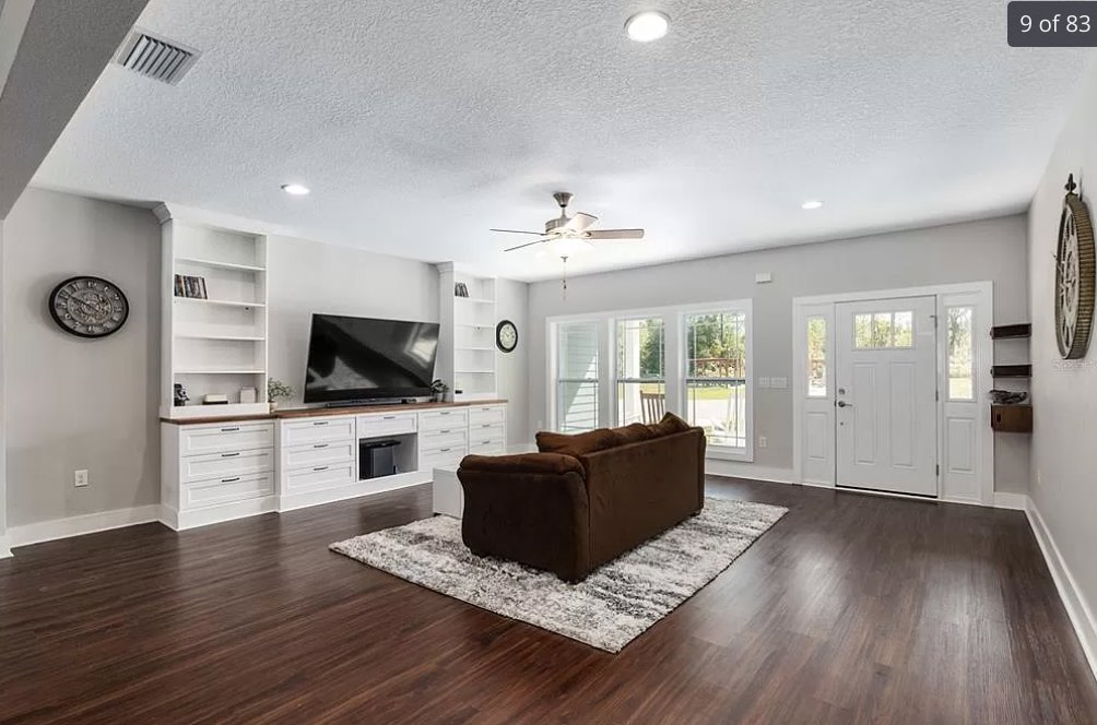 Brown upholstered couch facing white built-in cabinets, wood laminate flooring, patterned area rug, ceiling fan with light fixture, wall clock with roman numerals.