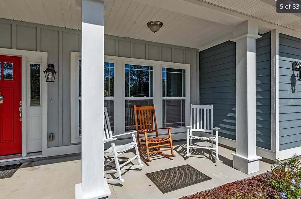 Cluster of wooden and white rocking chairs arranged on a spacious front porch with a black square mat, white flooring, paneled door, and ceiling light fixture.