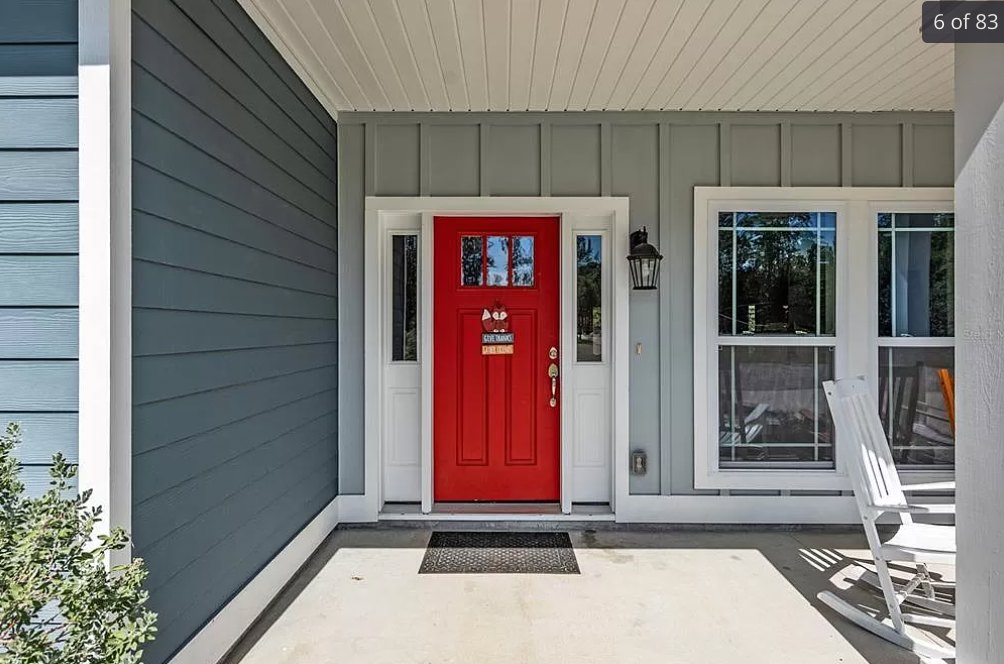 Red front door with decorative sign, white porch chair, stone siding, leafy tree partially obscuring blue exterior.