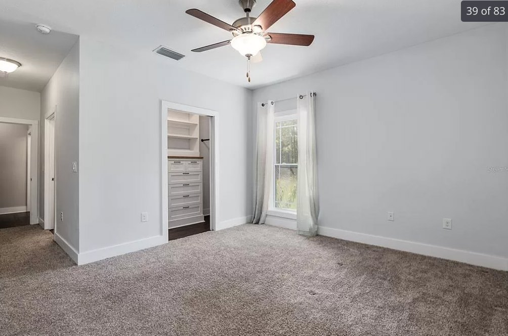 Carpeted room featuring a ceiling fan with light fixture, window with white curtains and frame, white walls, and plaster ceiling.