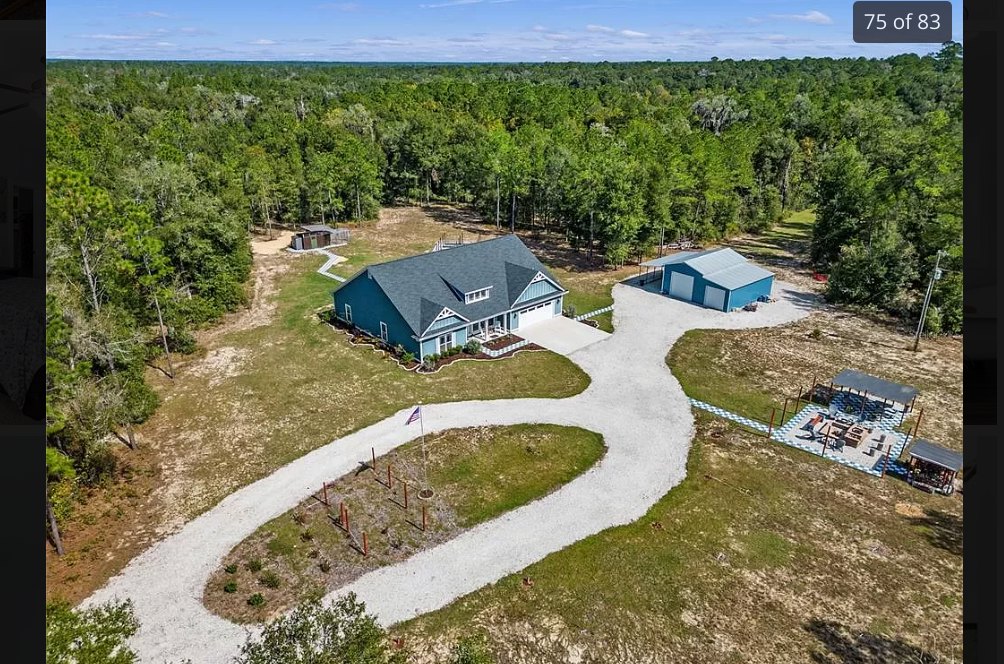 Blue-roofed house with white siding, concrete driveway, manicured lawn, mature trees, and partly cloudy sky