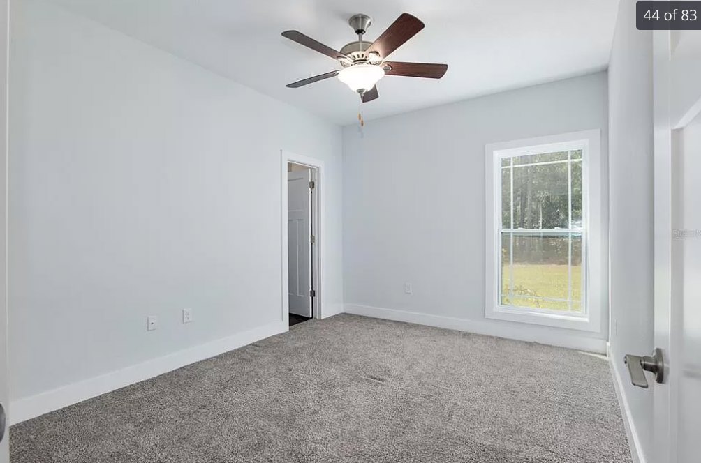 Bedroom with beige carpet flooring, white walls, ceiling fan with light fixture, white-framed window, and white door.