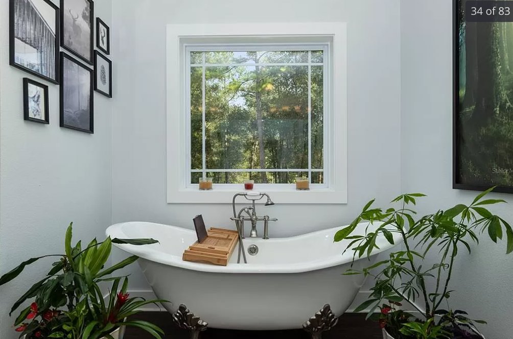 Freestanding bathtub with wooden tray holding a book, potted plants beside tub and toilet, large window overlooking trees, white walls and modern fixtures